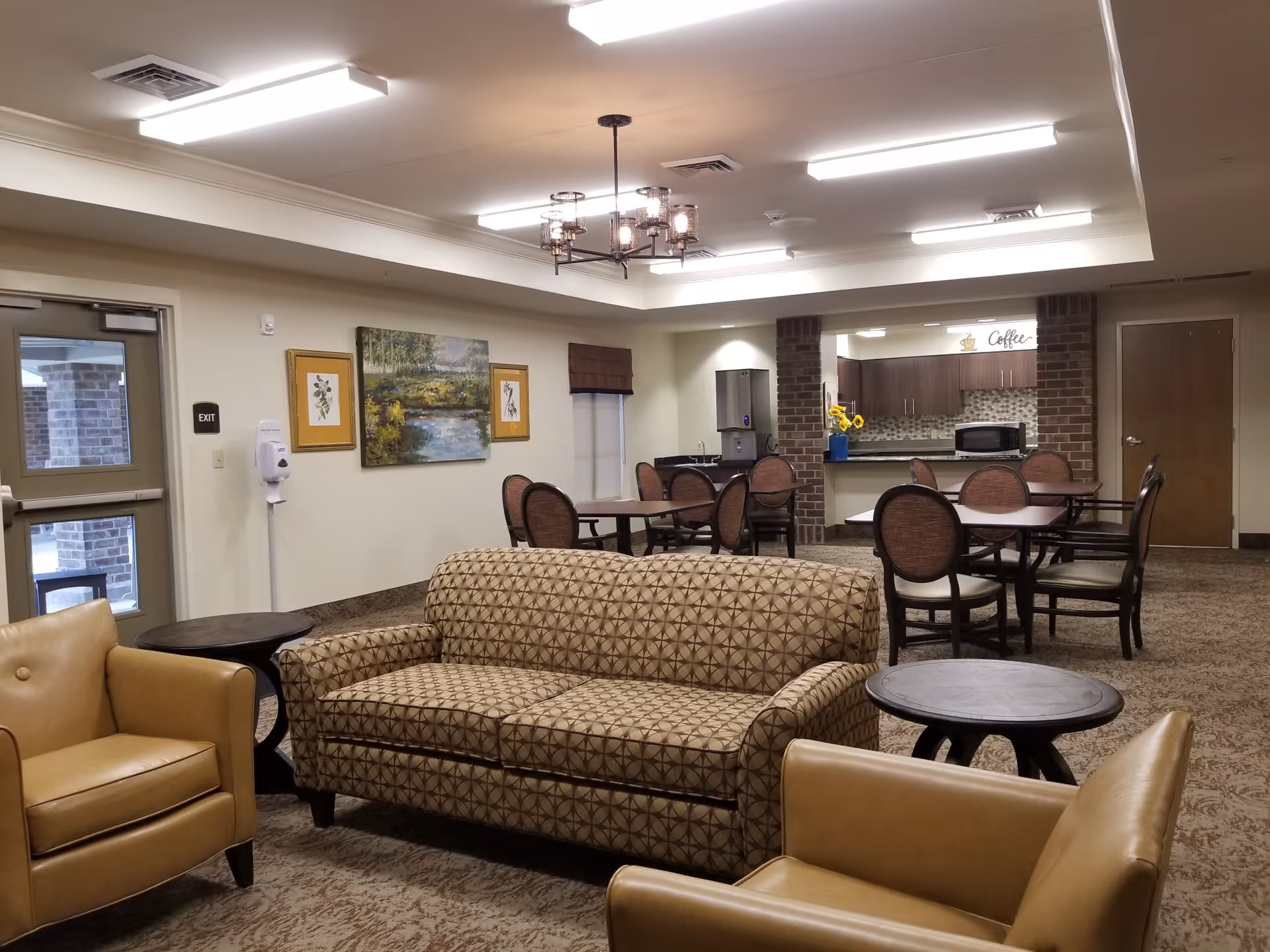 A cozy common area in a senior living facility featuring a patterned sofa and two tan armchairs arranged around two small round tables. In the background, there are several dining tables with chairs, a kitchenette area with a microwave, coffee station, and a water dispenser. The walls are decorated with framed artwork, and the ceiling has recessed lighting and a chandelier.