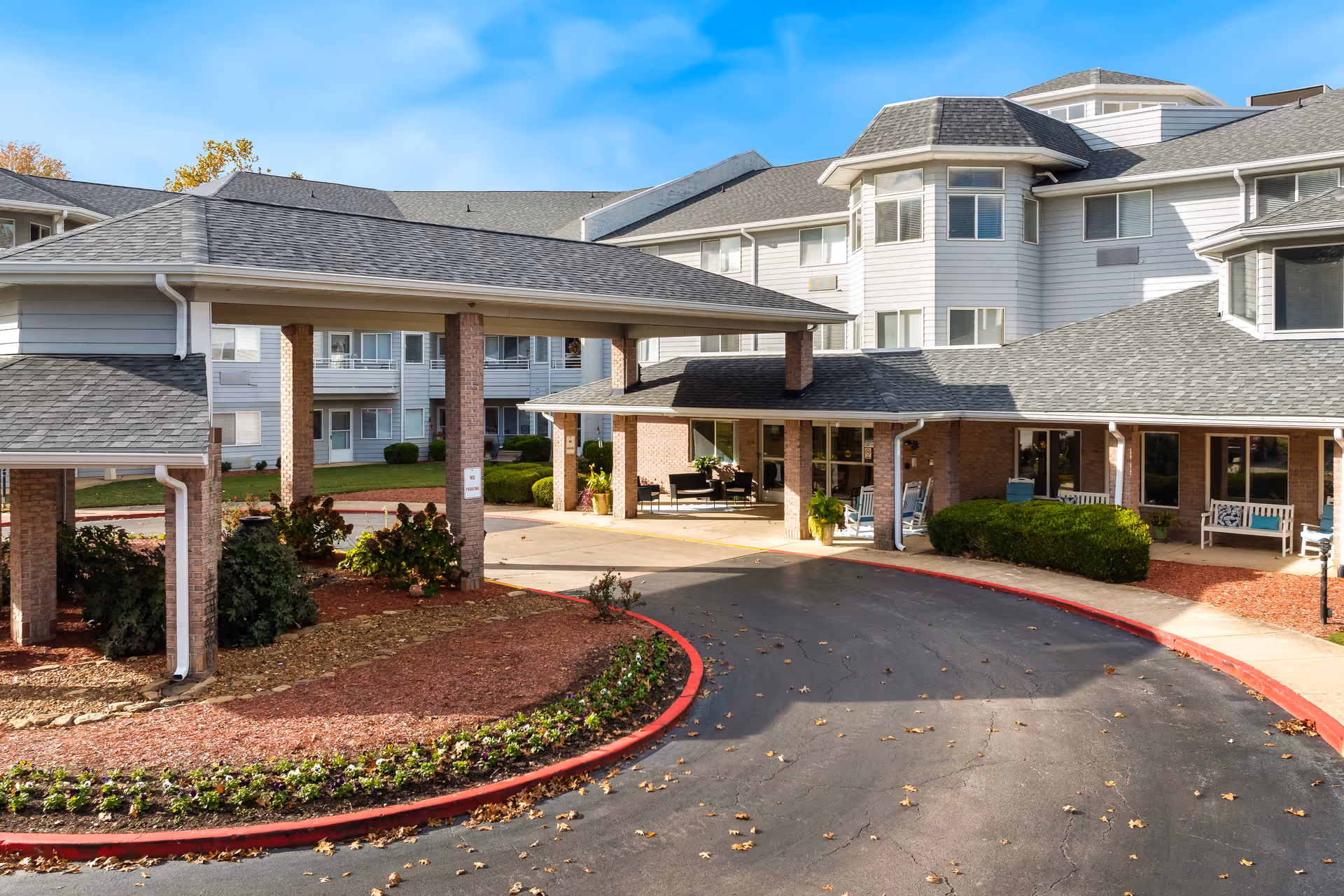 Exterior view of Jackson Meadow - A Provincial Senior Living Community, showing the main entrance with a covered drop-off area, surrounded by a curved driveway and landscaped garden beds with shrubs and flowers. The building is multi-story with white siding and multiple windows under a clear blue sky.