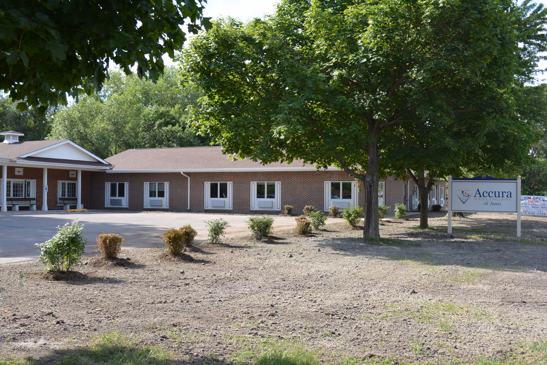 Exterior view of a single-story brick building with multiple windows and a covered entrance. There are several small bushes planted in front of the building and a large tree providing shade. A sign near the tree reads 'Accura HealthCare of Ames'.