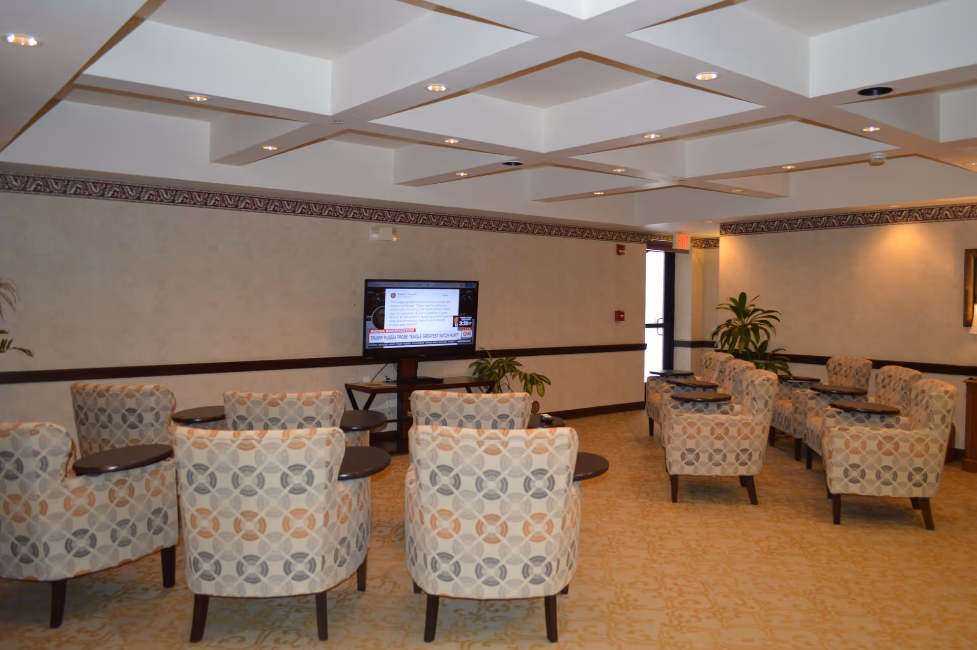 A senior living common room with patterned armchairs arranged facing a wall-mounted TV and small side tables.