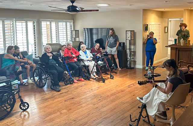 Elderly residents in wheelchairs and chairs sit in a common living room watching a woman play guitar.