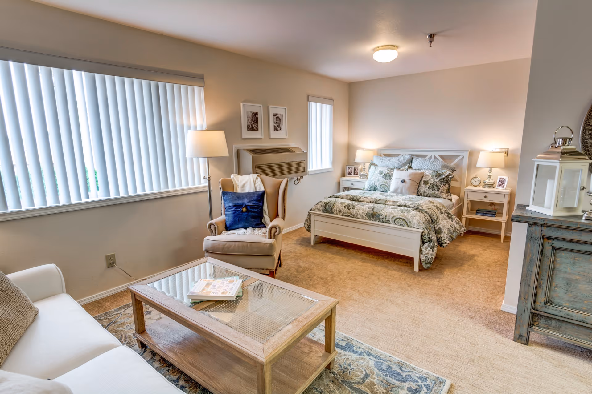 A cozy, well-lit room featuring a bed with patterned bedding, two bedside tables with lamps, a comfortable armchair with a blue pillow, a white sofa, and a wooden coffee table with books on it. The room has beige carpeting, vertical blinds on the windows, and neutral-colored walls.