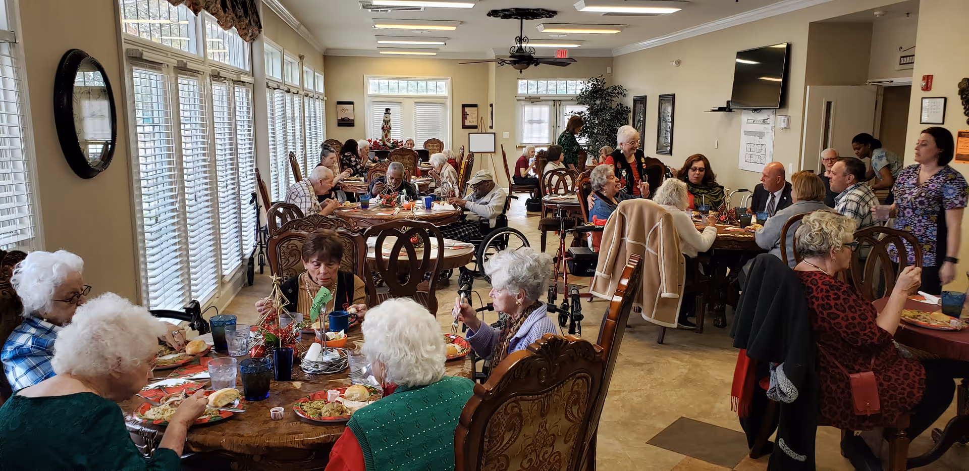 A large dining room where many elderly residents sit at tables eating and talking while staff circulate.