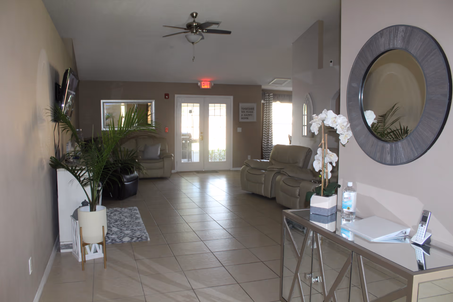 A bright and clean living room area with beige walls and tiled floor. The room features a ceiling fan, two beige recliner chairs, a large round mirror on the right wall, a glass console table with a white orchid plant, hand sanitizer, a phone, and a binder. There is a potted plant on the left side near a TV mounted on the wall. Double glass doors with an exit sign above them are visible at the far end of the room.