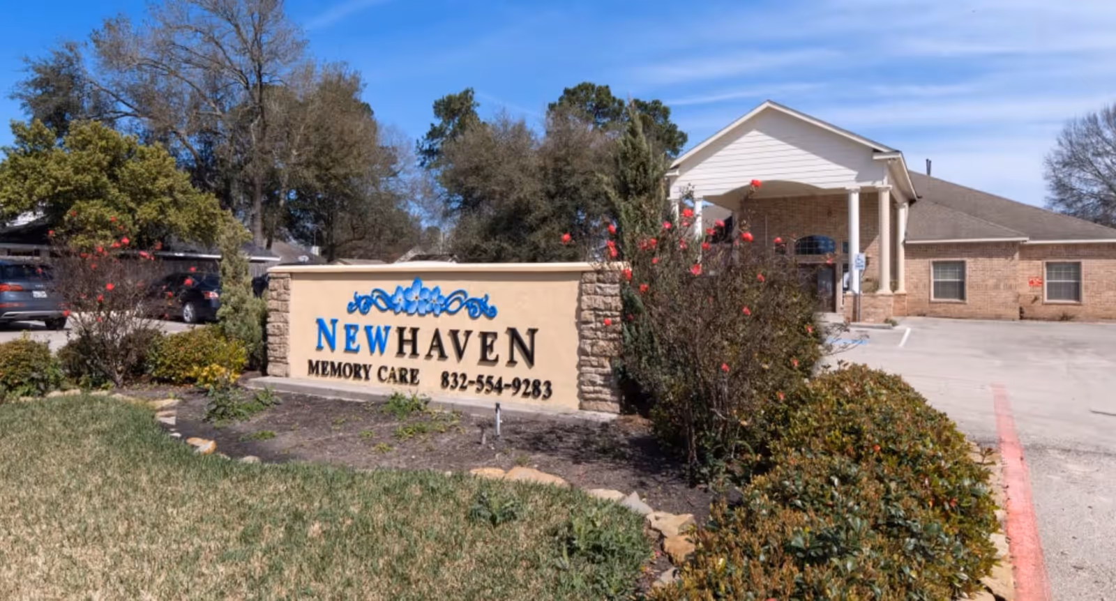 Stone entrance sign reading "NEW HAVEN MEMORY CARE" with the facility building, driveway, and landscaped shrubs under a blue sky.