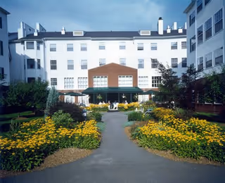 Outdoor garden area with a paved walkway leading to a white multi-story building with many windows. The garden is landscaped with yellow flowers and green shrubs, and there is a covered patio area with tables and chairs.