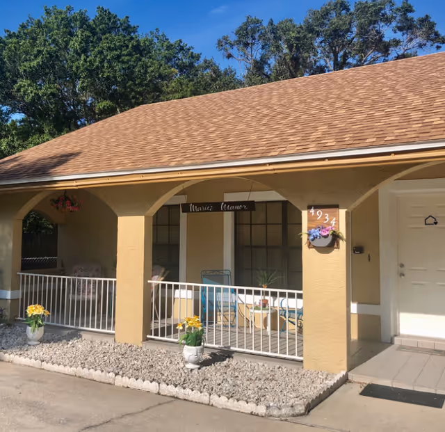 Exterior view of a single-story building with a tan stucco finish and a brown shingle roof. The building has a covered porch with white railings, two blue chairs, a small table, and potted plants. A sign hanging from the porch ceiling reads 'Marie's Manor'. The building number '4934' is displayed on a plaque with flowers next to the white front door. Trees and a clear blue sky are visible in the background.