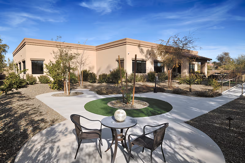 Outdoor courtyard with a round table and two chairs in front of a single-story beige building with landscaped paths under a blue sky.