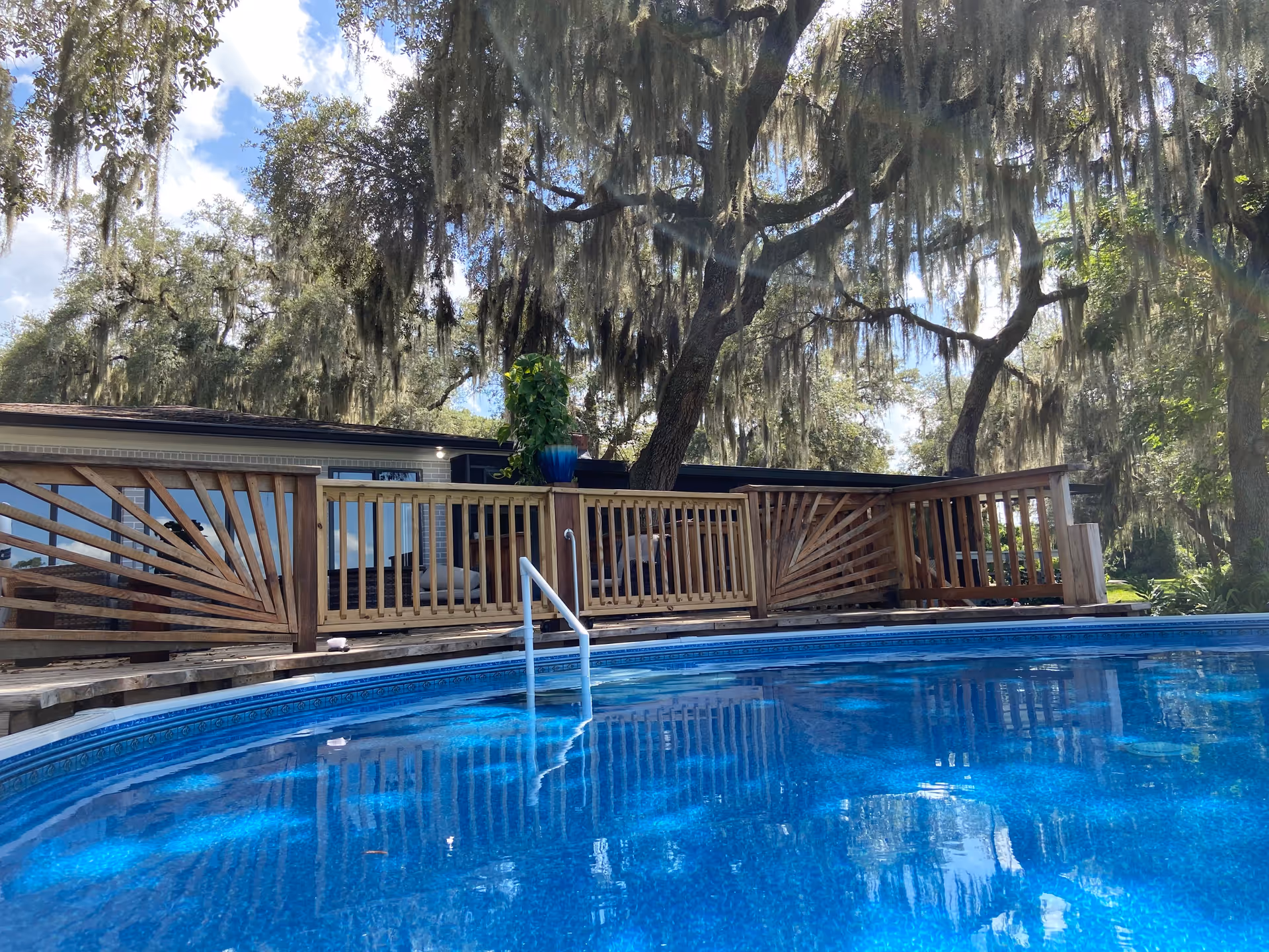 Outdoor view of a round above-ground swimming pool with clear blue water, surrounded by a wooden deck railing. Behind the pool, there are large trees draped with Spanish moss and a building partially visible under a partly cloudy sky.