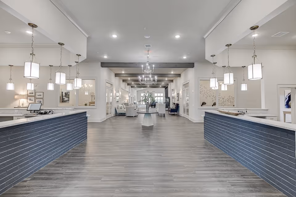 Spacious and modern reception area of Auburn Assisted Living with two blue brick front desks on either side, pendant lights hanging from the ceiling, and a seating area with chairs and a table in the background under exposed wooden beams.