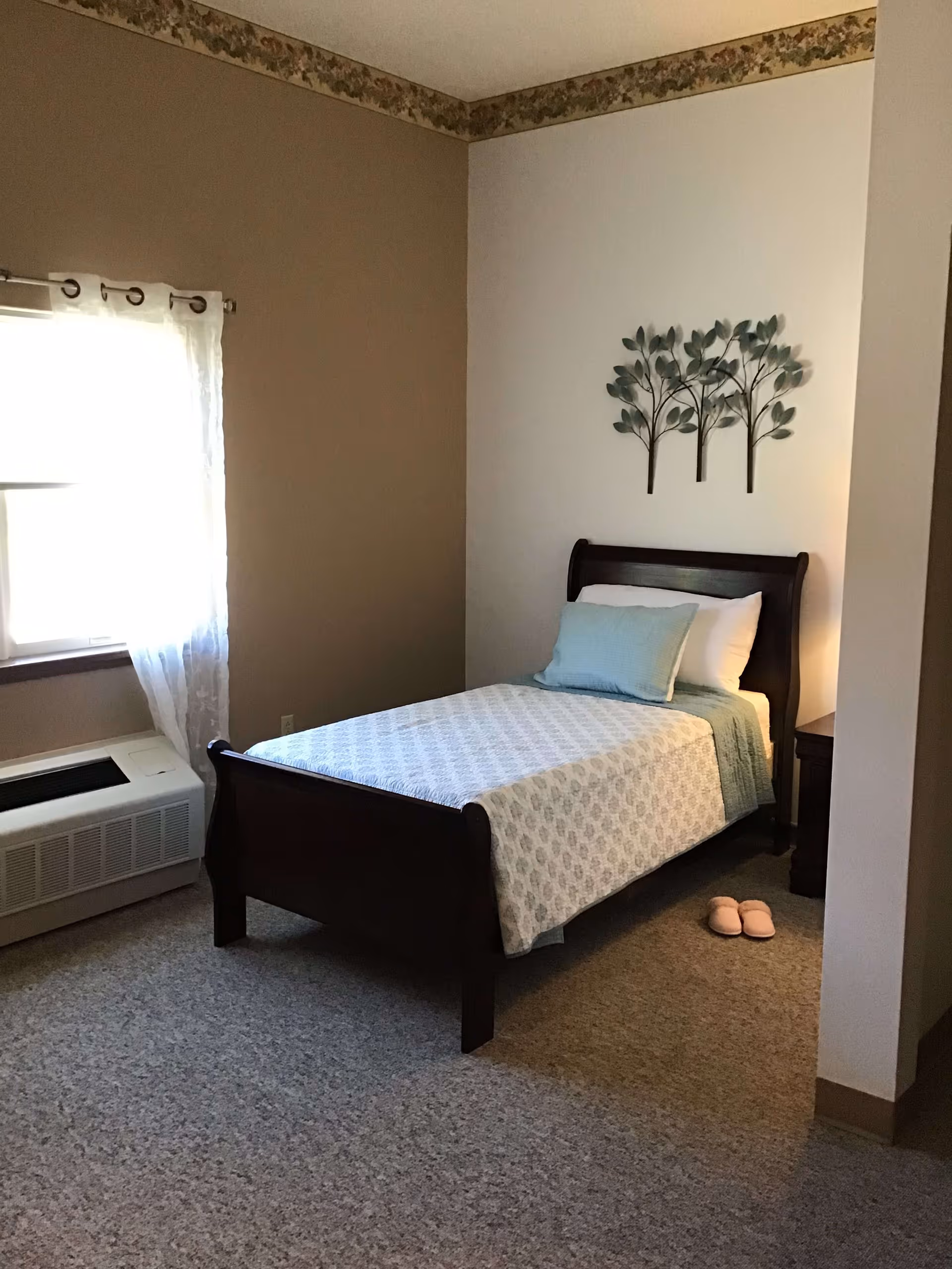 A small, cozy bedroom with a single dark wooden bed frame, a white and light blue patterned bedspread, and a light blue pillow. There is a pair of pink slippers on the carpeted floor next to the bed. A window with sheer white curtains lets in natural light. Above the bed, there is a decorative wall art piece featuring three stylized trees. The walls are painted in two tones, beige and white, with a floral border near the ceiling.