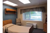 Small furnished bedroom with a single bed, wall shelf, chair, dresser, and a window overlooking greenery.