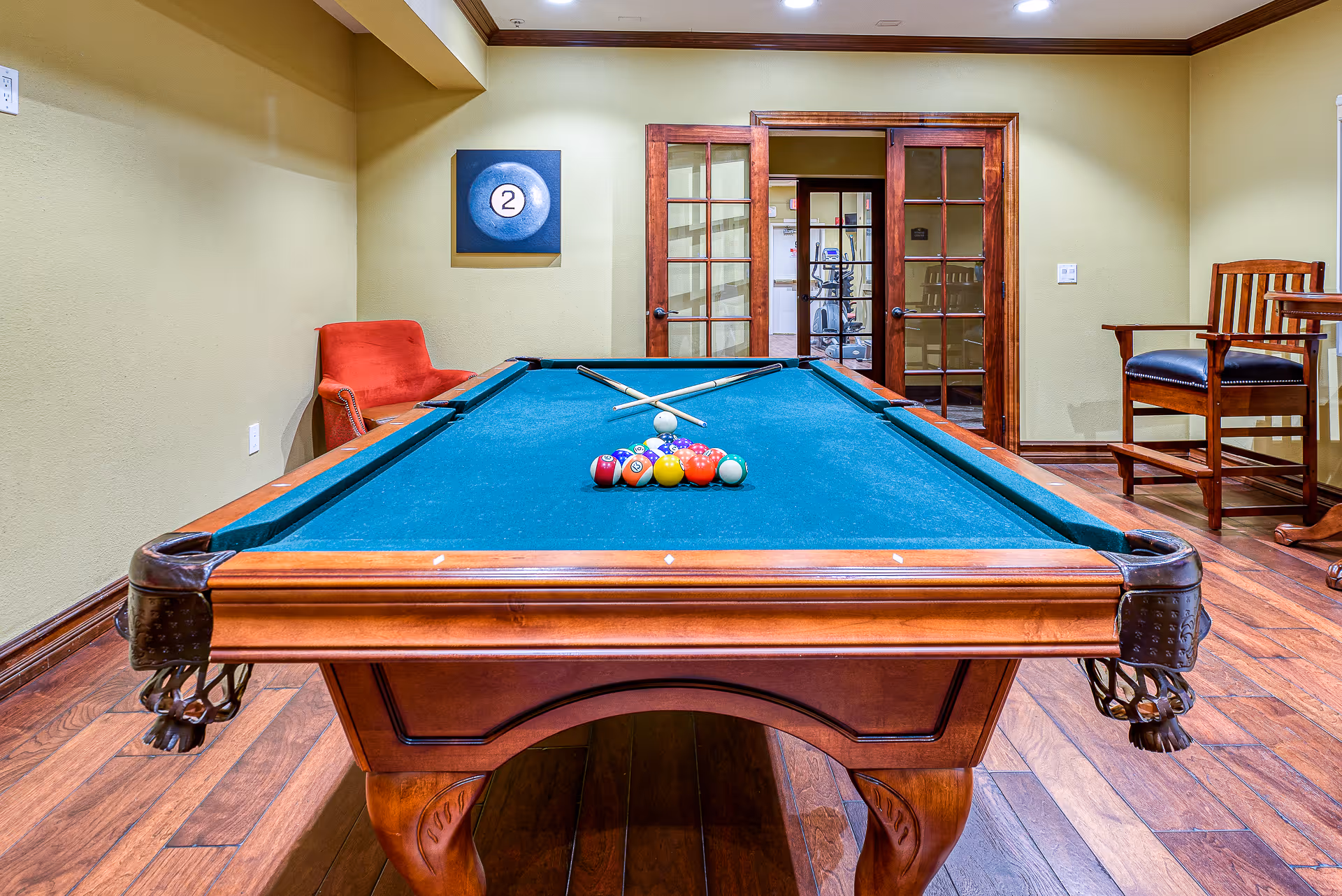 Interior room with a wooden pool table featuring a green felt surface, pool balls arranged in a triangle, and two crossed pool cues. The room has wooden flooring, beige walls, a red chair on the left, and wooden chairs and a table on the right. A framed picture of a billiard ball with the number 2 hangs on the wall. Glass double doors lead to another room with exercise equipment.