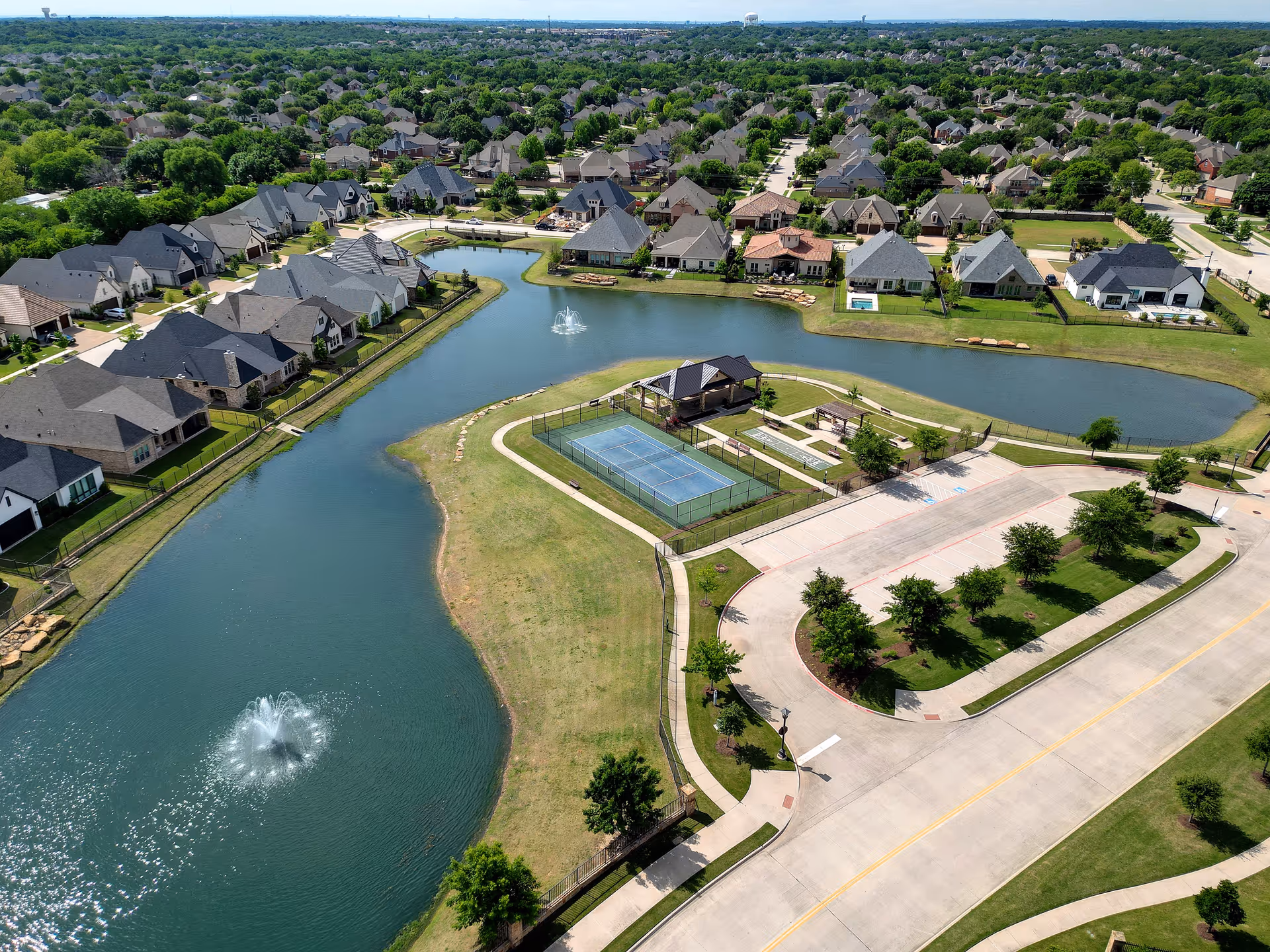 Aerial view of a residential neighborhood with a water feature including two fountains, a tennis court, a small pavilion, and surrounding houses with well-maintained lawns and streets.
