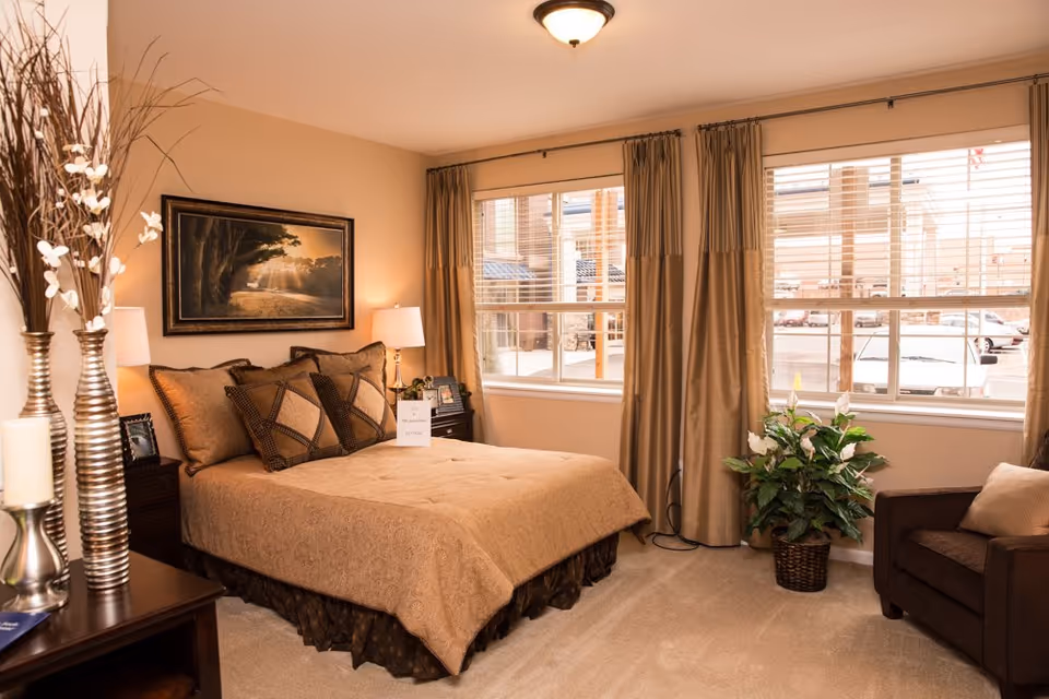 A warmly decorated bedroom with a large bed covered in a beige bedspread and multiple pillows. The room features two windows with beige curtains, a brown armchair with a beige pillow, a potted plant, and a nightstand with a lamp and framed photos. There is a framed landscape painting on the wall above the bed and decorative vases with white flowers on a dresser.