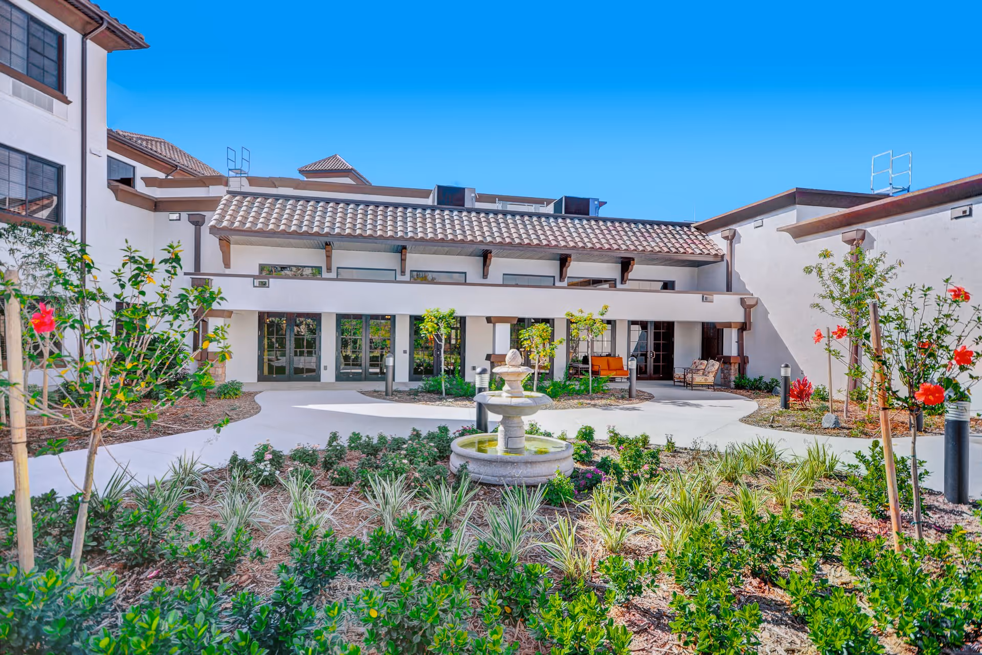 Sunny courtyard with a central fountain, landscaped plant beds and seating in front of a Mediterranean-style building.