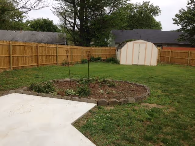 A fenced backyard with a small garden bed bordered by stones, a white shed in the background, and a concrete patio in the foreground. Trees and neighboring houses are visible beyond the wooden fence.