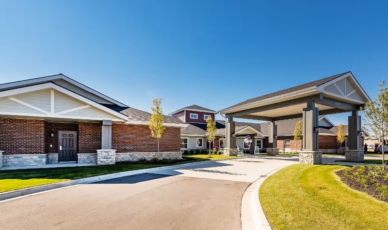 Front exterior of a senior living facility showing brick buildings and a covered entrance drive under a clear blue sky.