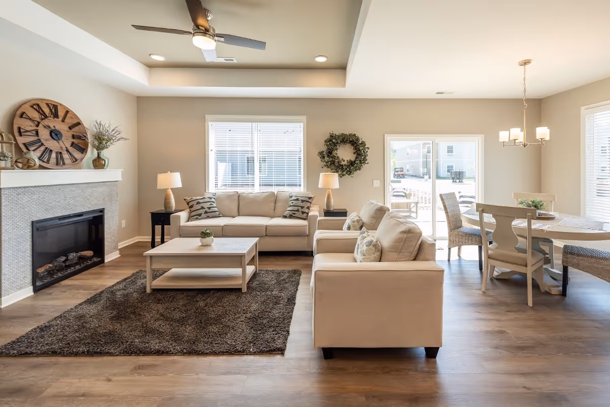 A bright and spacious living room with beige sofas, a wooden coffee table on a dark rug, a fireplace with a large decorative clock above it, two table lamps on side tables, and a dining area with a round table and chairs near sliding glass doors leading outside.