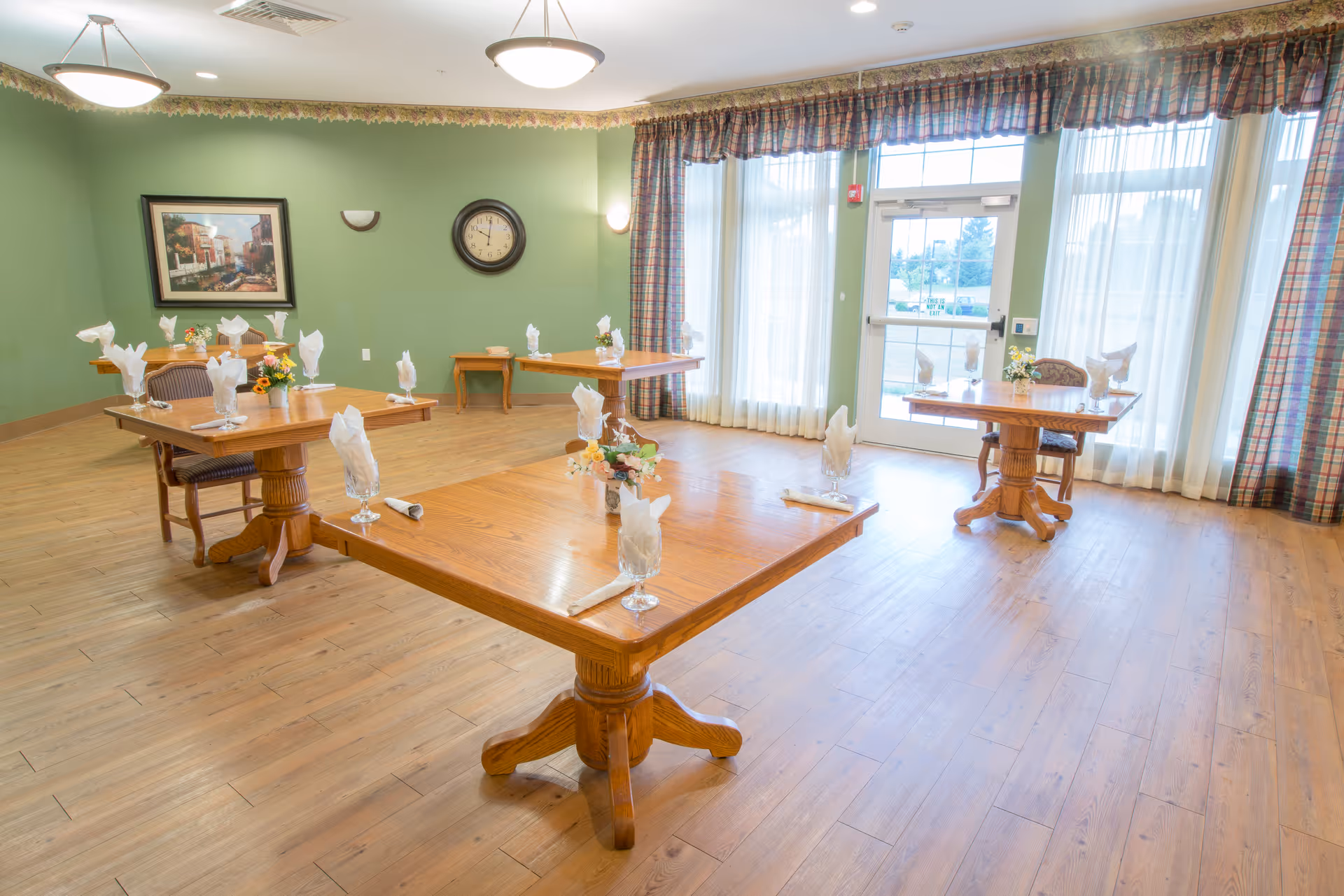 A bright dining room with wooden floors and green walls. There are four wooden tables, each set with folded white napkins in glasses and small floral centerpieces. The room has large windows with sheer white curtains and plaid valances, allowing natural light to fill the space. A clock and a framed picture hang on the wall.