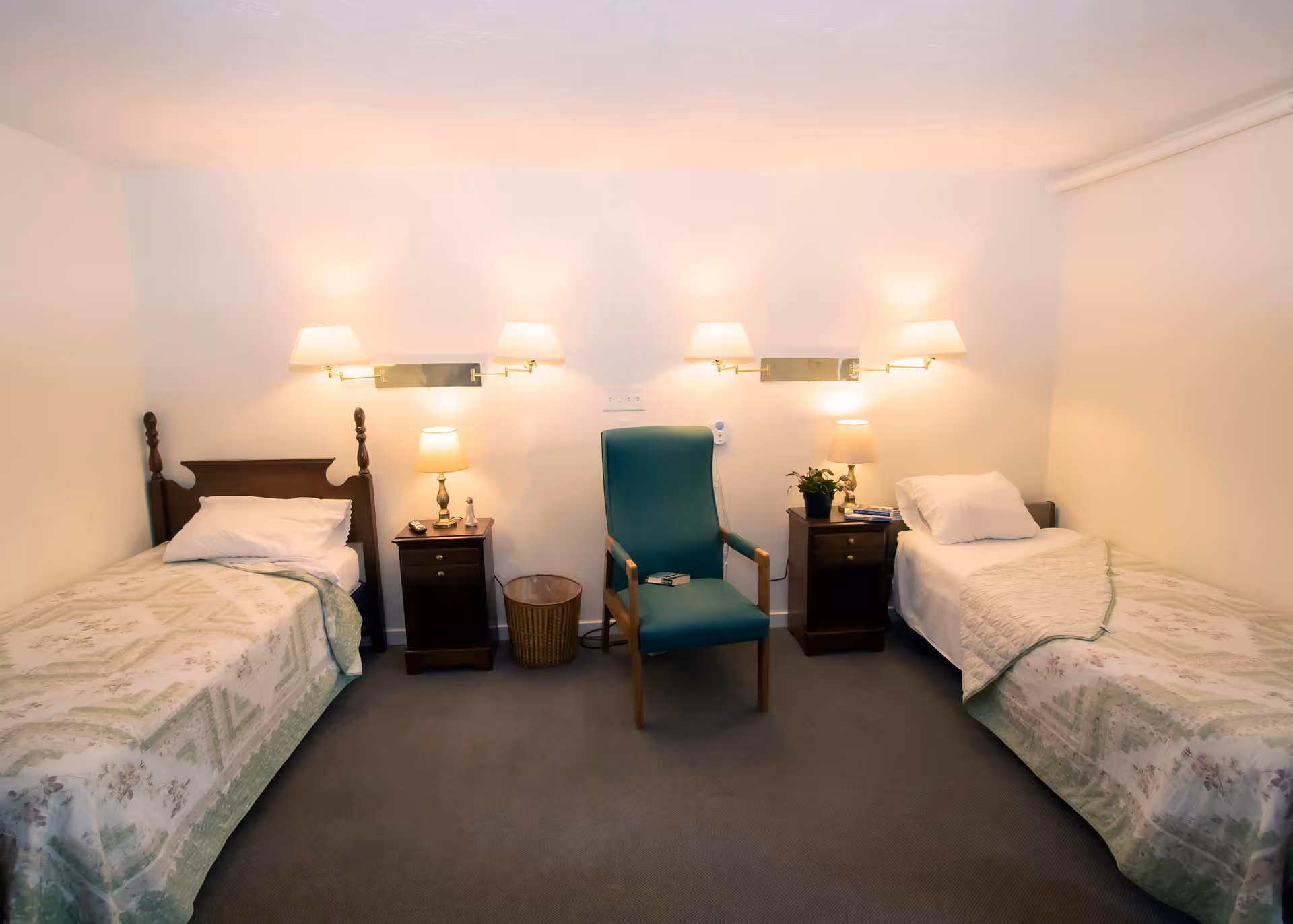 A simple bedroom with two single beds on either side of the room, each with a nightstand and lamp. A green armchair is positioned in the center between the beds. The walls are white, and there are wall-mounted lamps above each bed providing warm lighting.