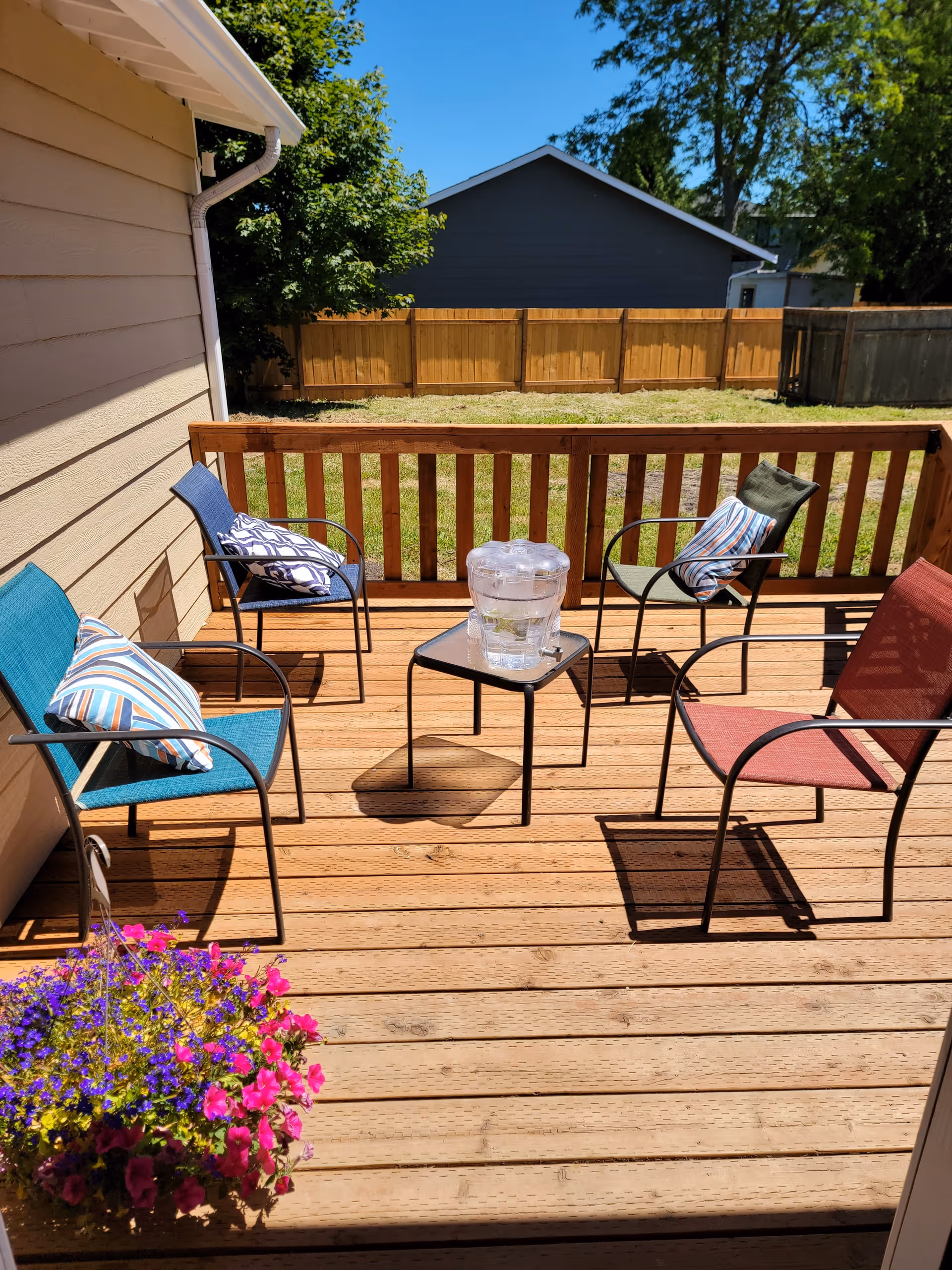 A wooden outdoor deck with four metal chairs, each with a colorful cushion. A small square table in the center holds a clear water dispenser. There is a vibrant flower pot with pink and purple flowers on the left side of the deck. The deck is enclosed by a wooden railing, and a fenced grassy yard with trees and a neighboring house is visible in the background under a clear blue sky.