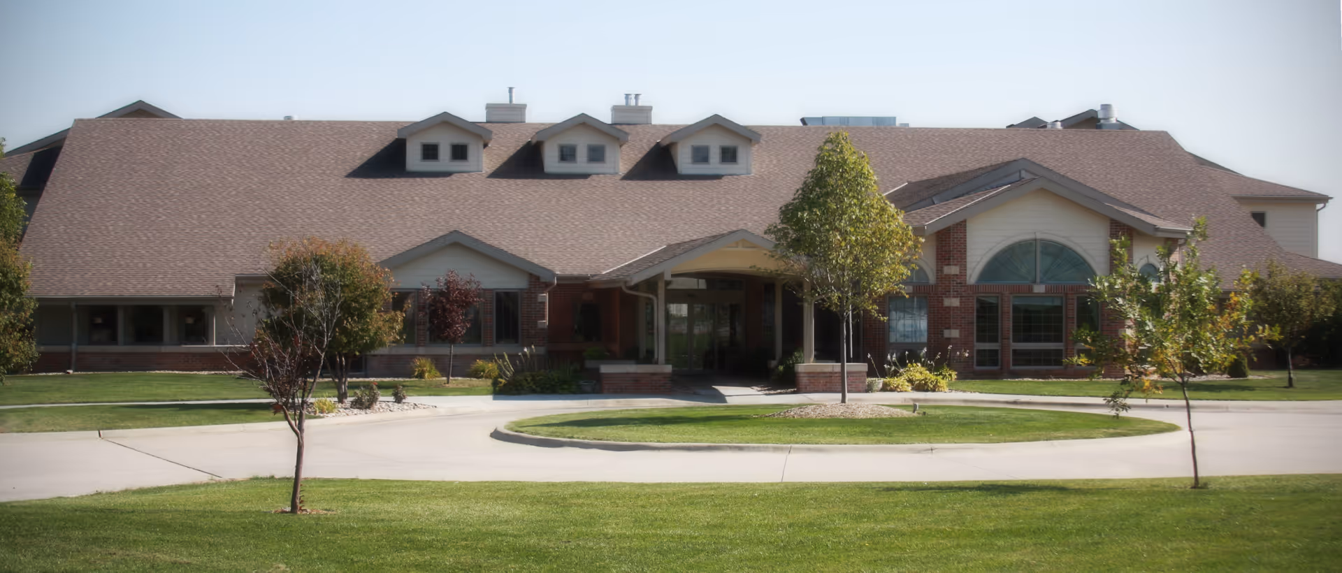 Front exterior of a single-story brick senior living facility with a covered entrance, dormer windows, circular driveway, and landscaped lawn.