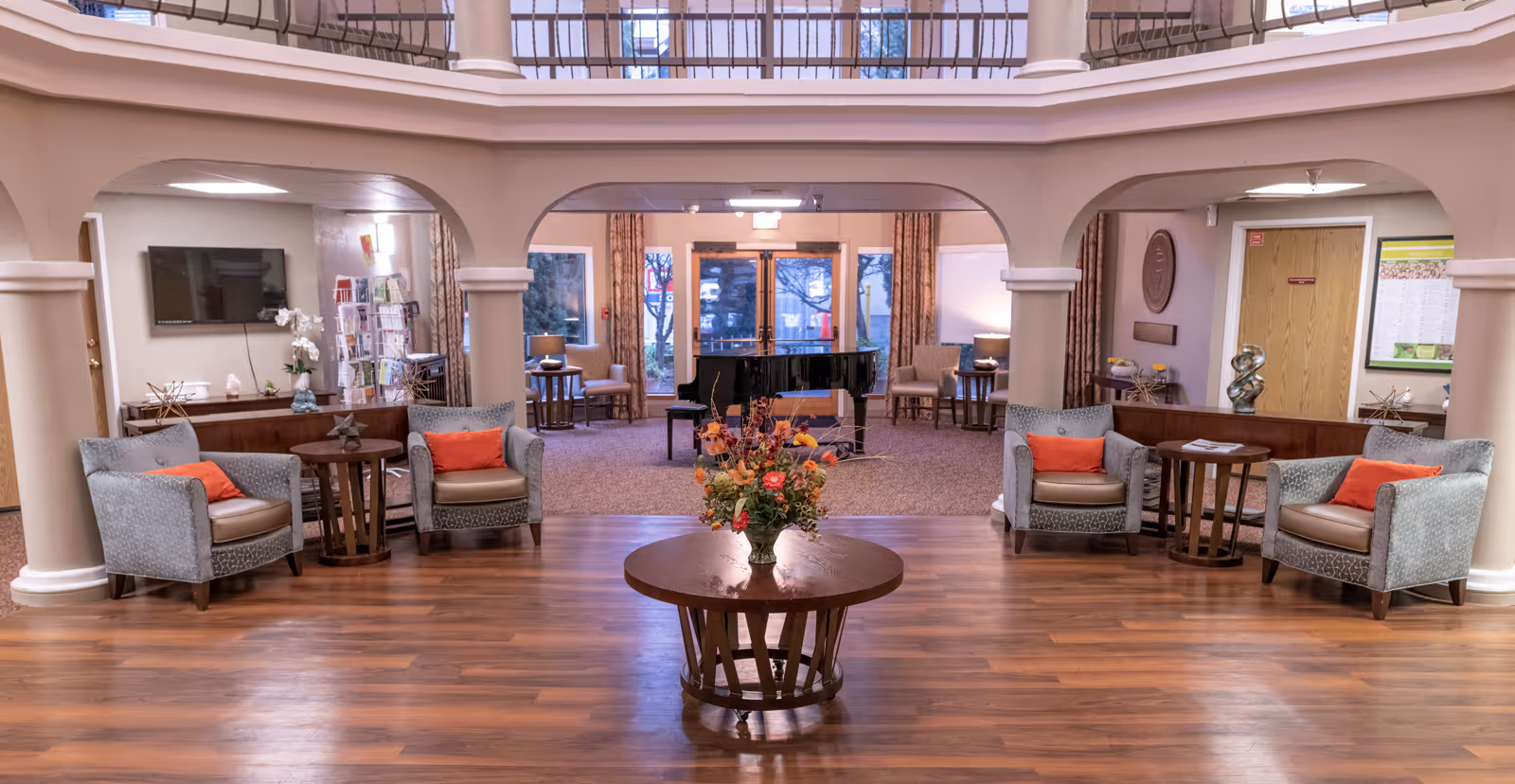 A spacious and well-lit senior living facility common area with wooden flooring and a round wooden table in the center holding a floral arrangement. The room features four armchairs with orange cushions arranged in pairs on either side of the table, small side tables, and a grand piano near the glass double doors at the back. The area is framed by large columns and arches, with a second-floor balcony visible above.
