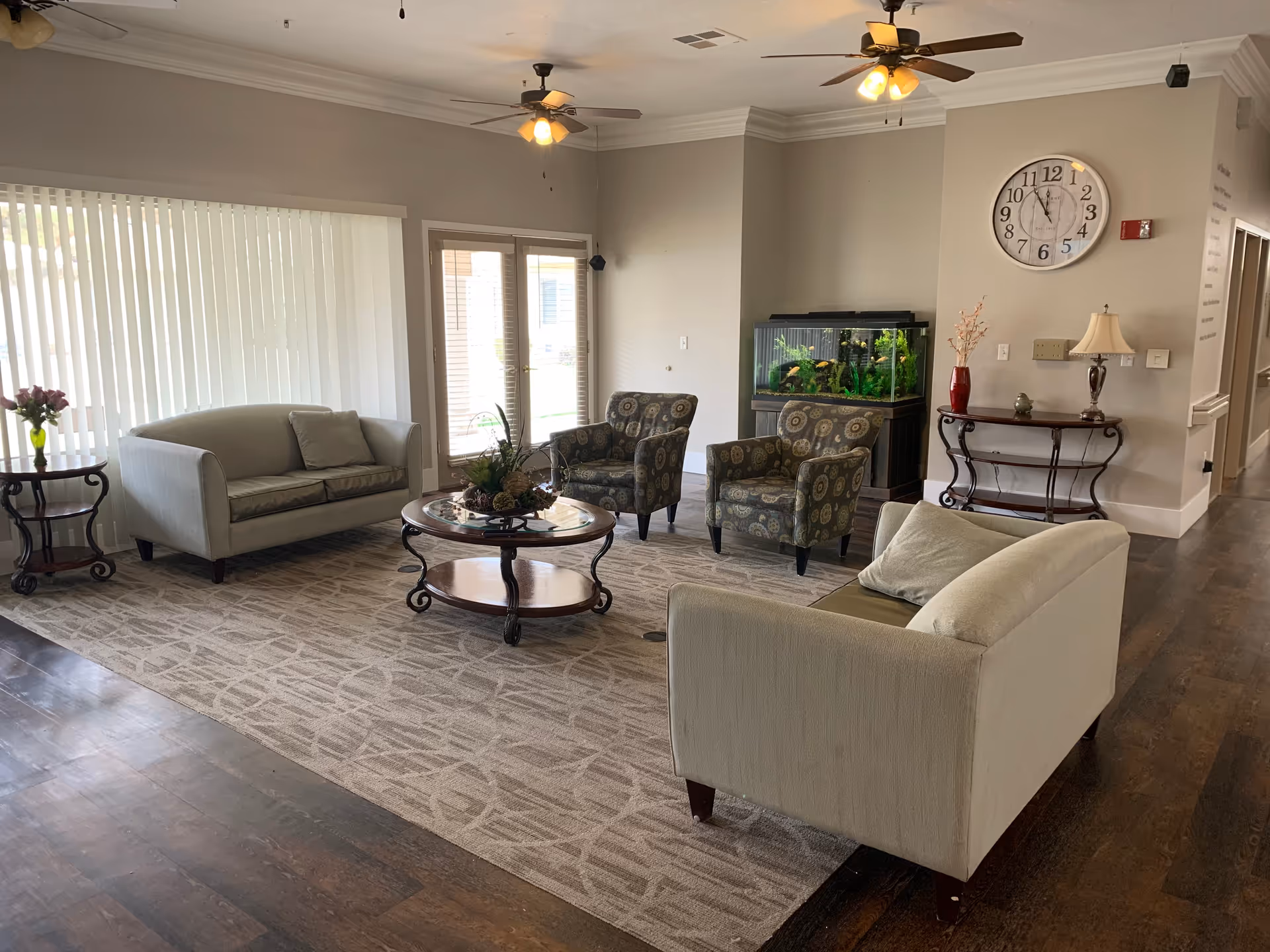 A cozy assisted living facility common area with two beige sofas, two patterned armchairs, a round glass coffee table with a floral centerpiece, and a side table with a vase of flowers. The room has large vertical blinds covering a window, two ceiling fans with lights, a wall clock, a fish tank, and a small console table with a lamp and decorative items. The floor is a combination of dark wood and a patterned carpet.
