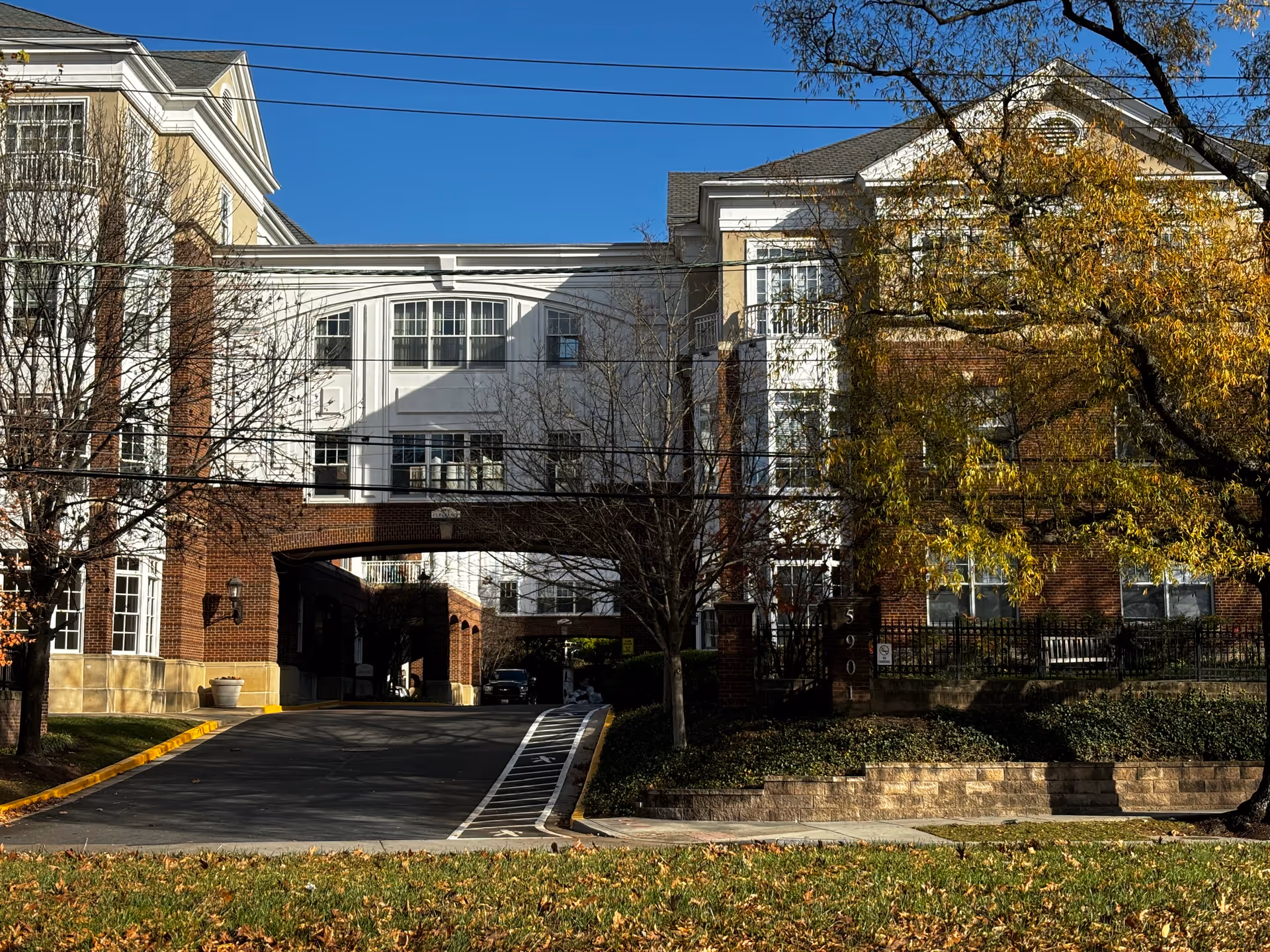 Exterior view of a multi-story assisted living community building with brick and white siding, featuring a covered driveway entrance and surrounded by trees with autumn foliage under a clear blue sky.