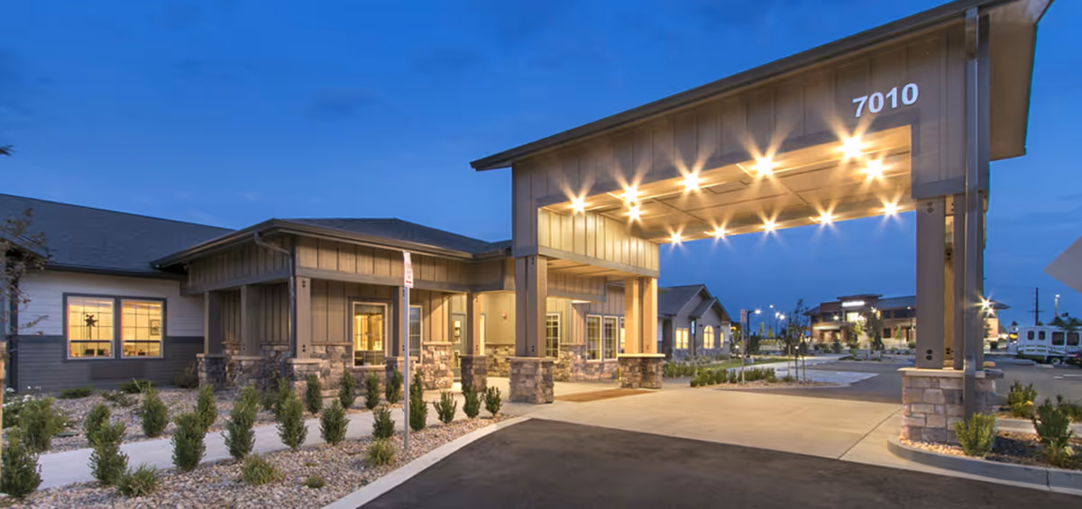 Exterior view of a senior living facility entrance at dusk with a covered drop-off area illuminated by multiple lights. The building features stone and wood siding with large windows, and the address number 7010 is displayed prominently on the canopy. Landscaping with small shrubs lines the walkway leading to the entrance.