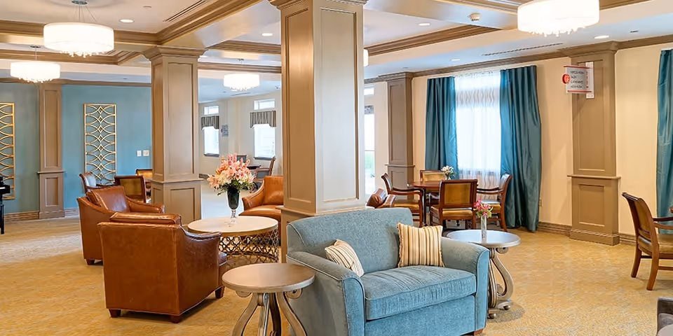 A spacious and well-lit living room area in a senior living facility featuring comfortable seating including a blue loveseat with striped pillows, brown leather armchairs, round side tables, and a round table with a vase of flowers. The room has beige walls with decorative columns, teal curtains, and multiple chandeliers providing warm lighting.