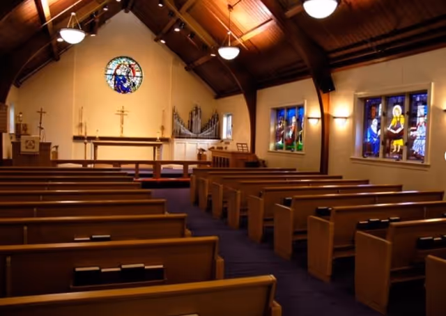 Interior of a chapel with wooden pews facing an altar, stained-glass windows, and a vaulted wooden ceiling.