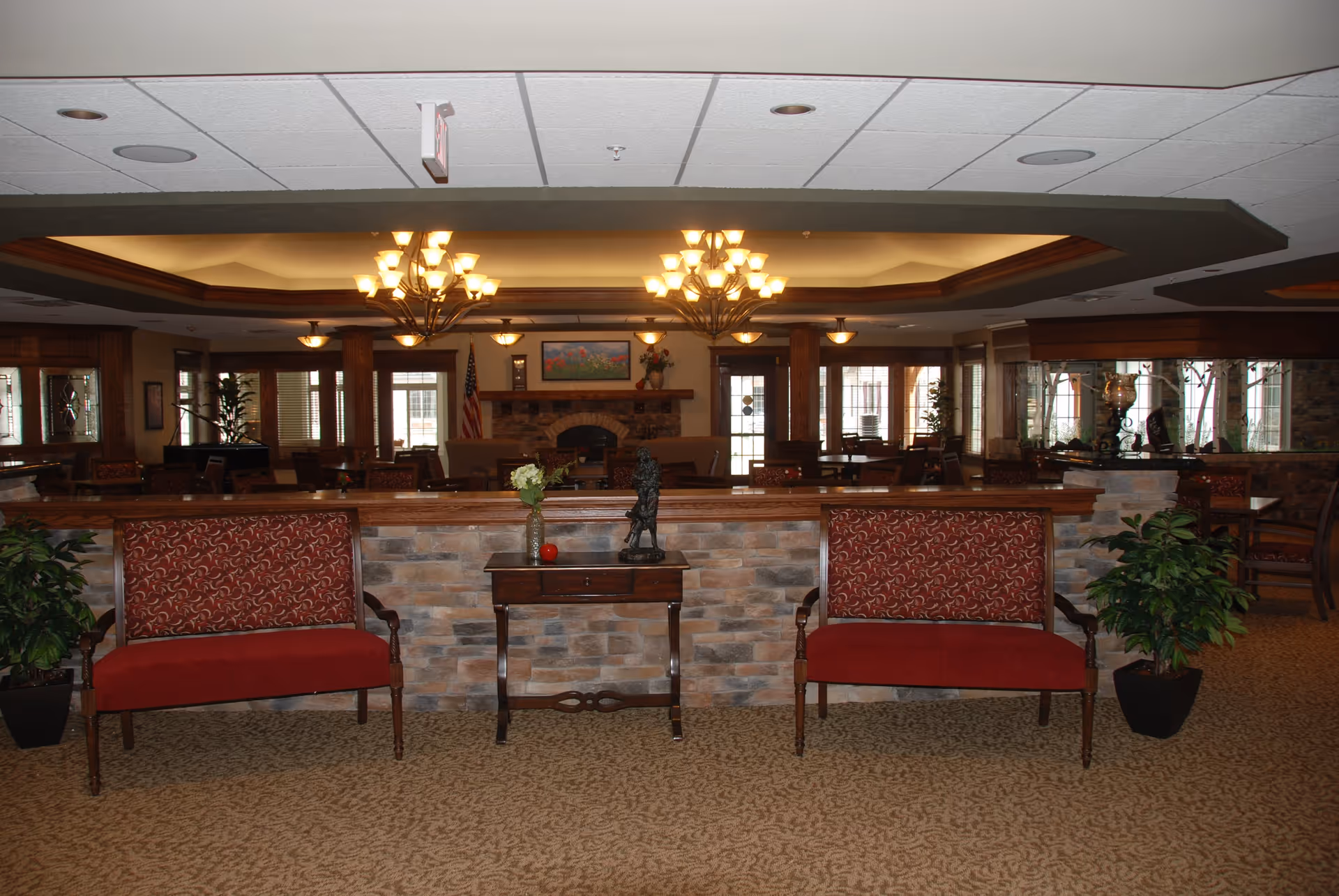 Interior view of a senior living facility lounge area with two red upholstered benches separated by a small wooden table holding a vase with flowers and a decorative statue. The background features a stone half-wall, multiple tables and chairs, large windows, an American flag, a fireplace, and warm ceiling lighting fixtures.