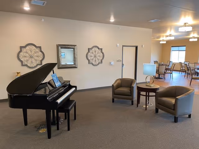 A spacious room with a black grand piano on the left side, two brown leather armchairs with nailhead trim arranged around a small round wooden table with a modern lamp on it. The walls are beige with two decorative wall clocks and a framed mirror. In the background, there is a dining area with tables and chairs near windows.