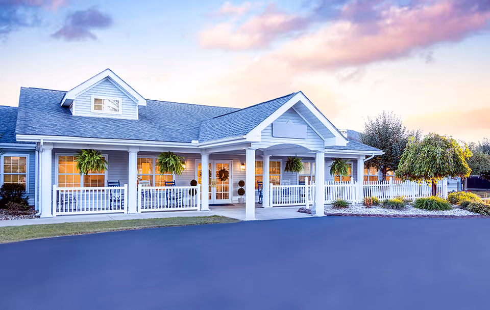Exterior view of a single-story building with a covered porch featuring white columns and railings. The porch has hanging plants and rocking chairs. The building is light blue with white trim, and there is a paved driveway in front. The sky is partly cloudy with a warm sunset glow.