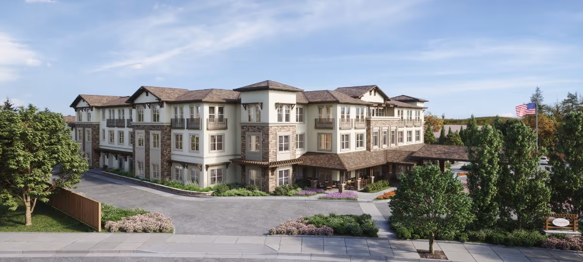 Front exterior of a three-story senior living building with landscaped grounds, a driveway, and an American flag.