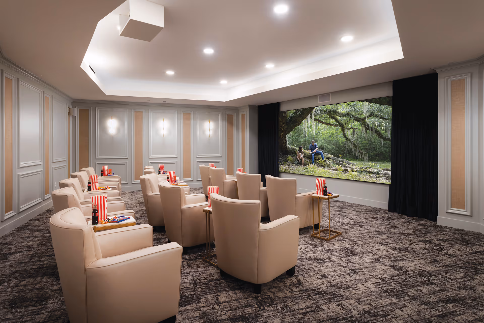 A cozy movie theater room with beige armchairs arranged in rows facing a large screen showing an outdoor scene with two people sitting under a tree. Each chair has a small side table holding popcorn, candy, and a soda bottle. The room has soft lighting, paneled walls, and a carpeted floor.