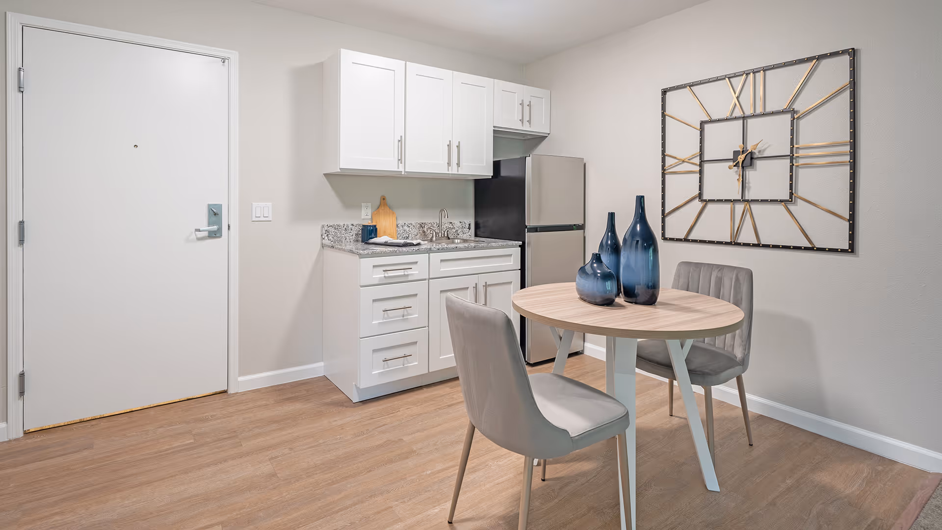 Compact kitchenette and dining nook with white cabinets, a small stainless refrigerator, a round wood table with two chairs, and a large decorative wall clock.