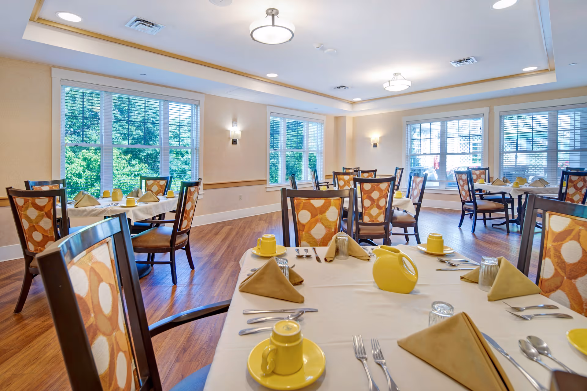 Bright dining room with multiple tables set with yellow cups, beige napkins, silverware, and yellow pitchers. The room has large windows with white blinds letting in natural light, wooden floors, and beige walls with modern light fixtures.