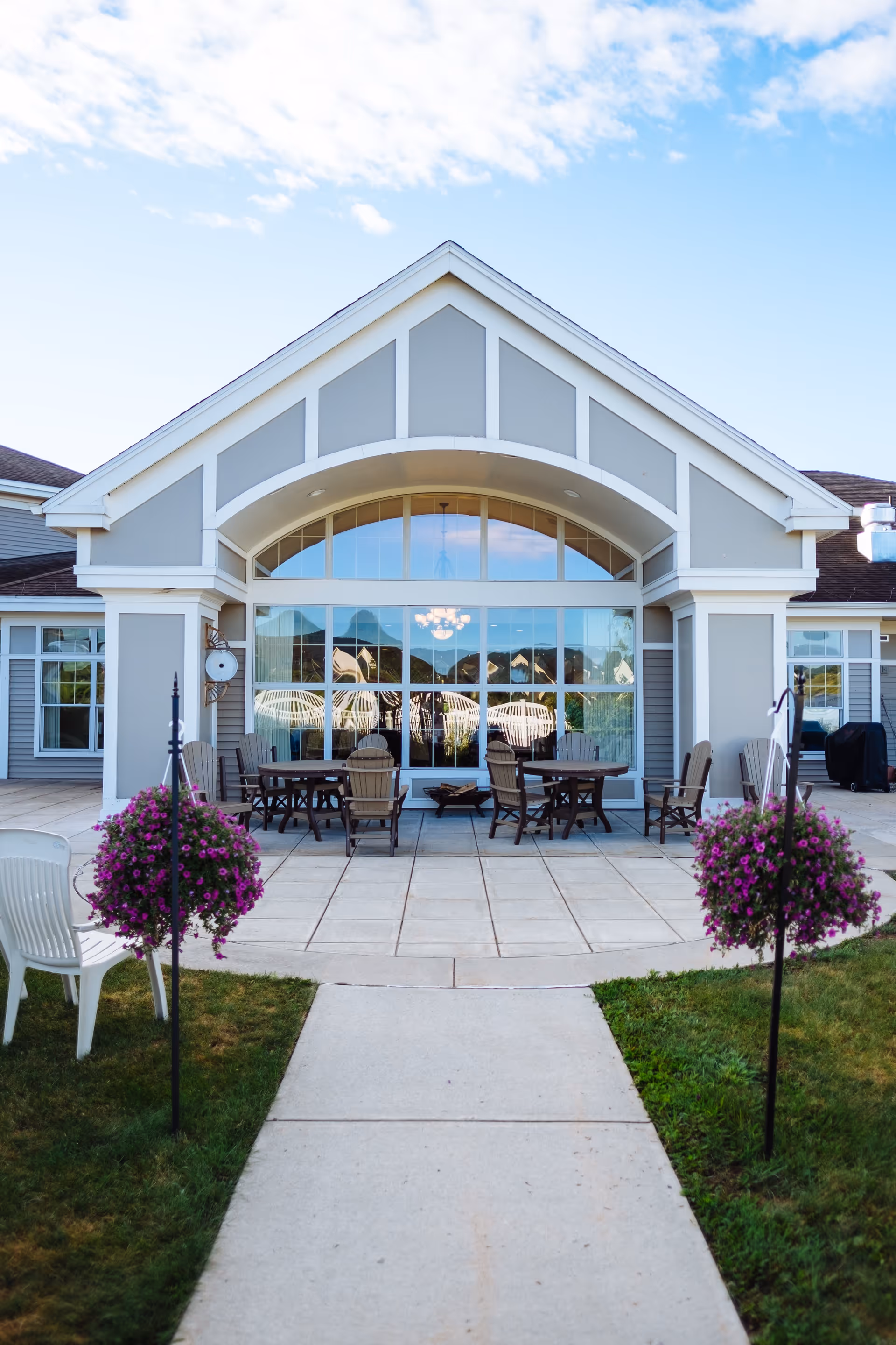 Outdoor patio area at The Terrace at The Glen with a paved walkway leading to a seating area with tables and chairs. The building has large windows and a peaked roof, with hanging flower baskets on either side of the walkway.