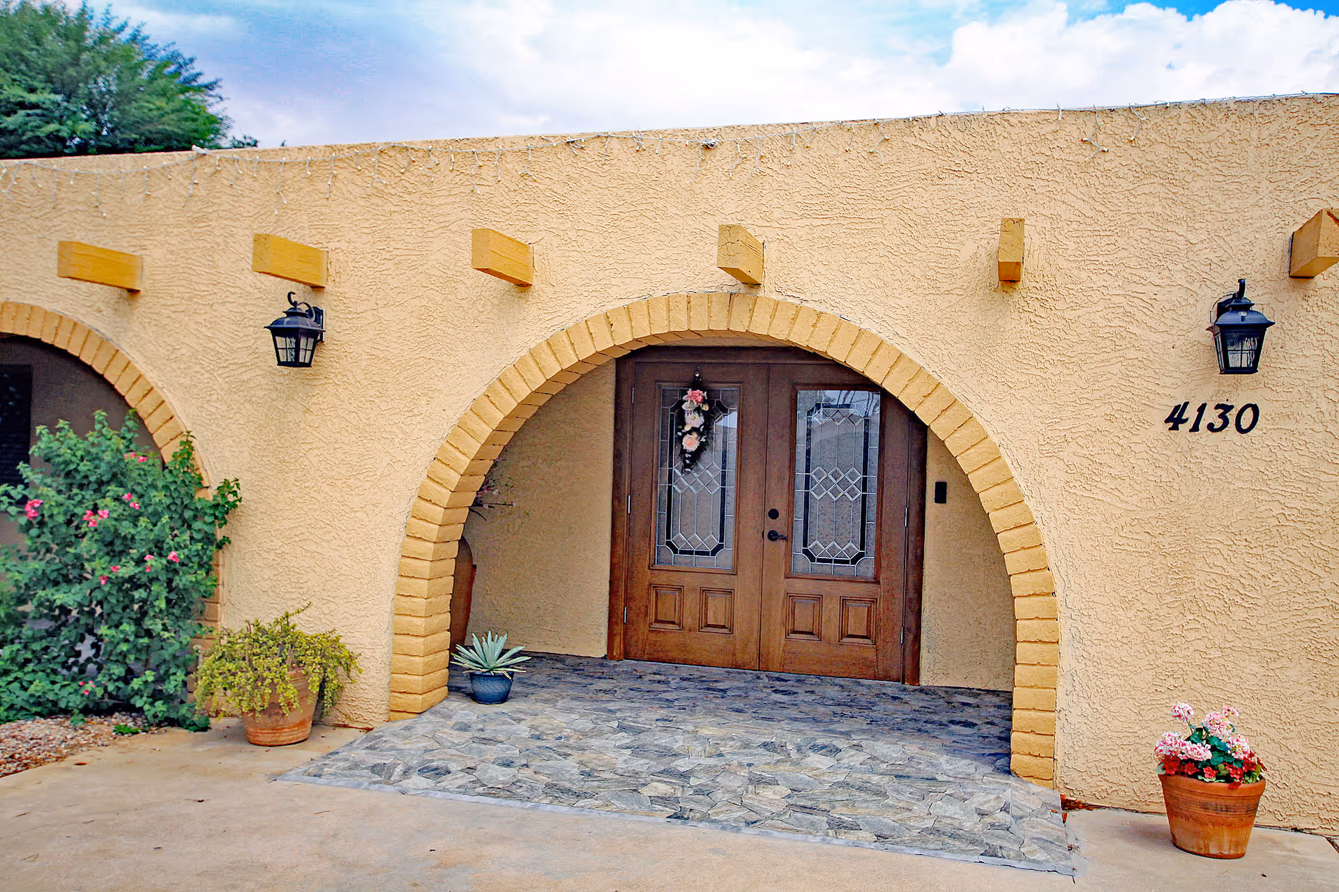 Exterior view of a tan stucco building entrance with arched doorway and potted plants.