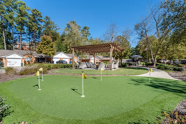 Small outdoor putting green with flags in front of a pergola-covered seating area, surrounded by garages, trees, and walkways.