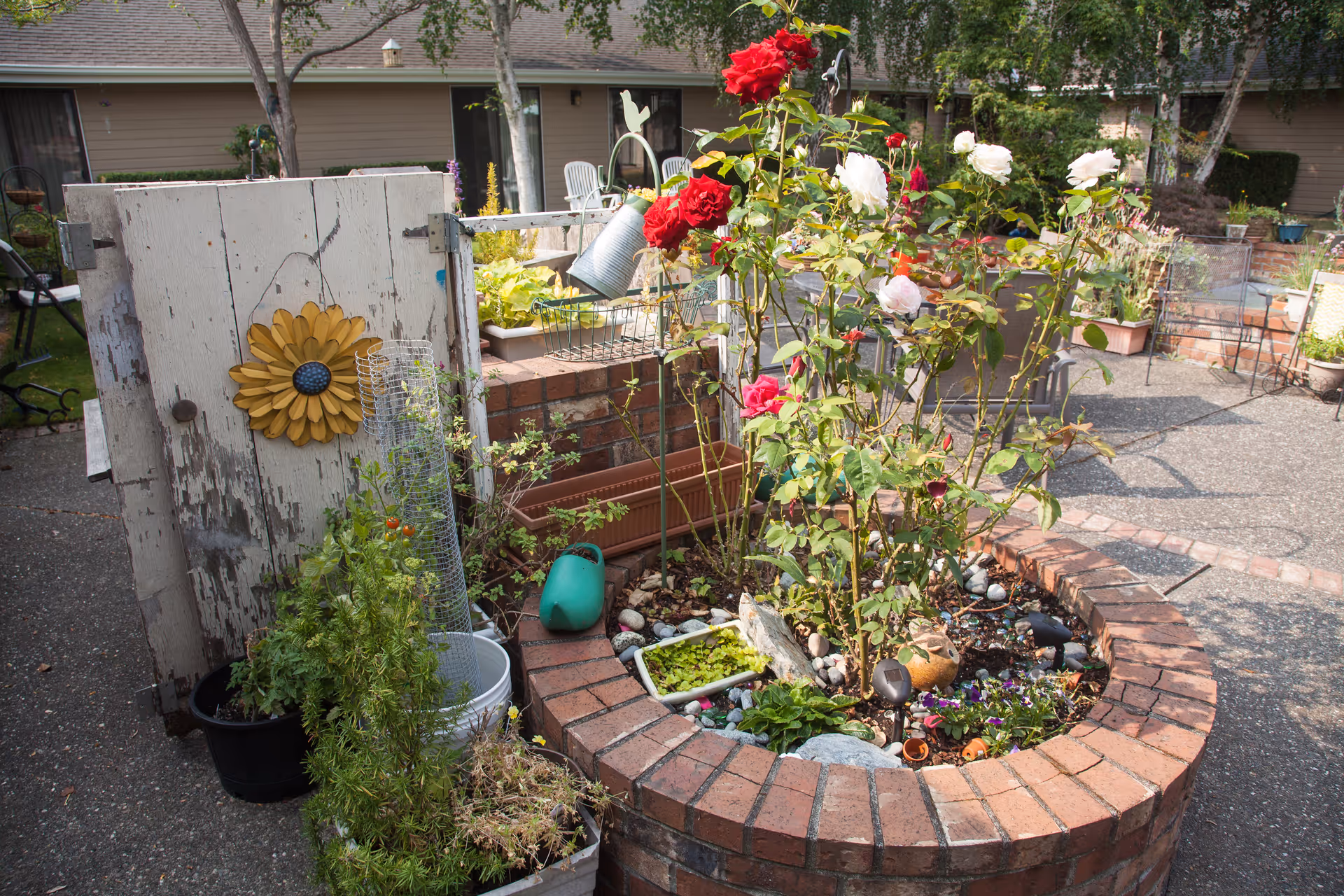 A circular brick planter filled with blooming red and white roses, small plants, and decorative items. Surrounding the planter are various gardening tools and pots, including a watering can and a rustic wooden door with a yellow flower decoration. In the background, there are outdoor chairs and a building with windows and trees.