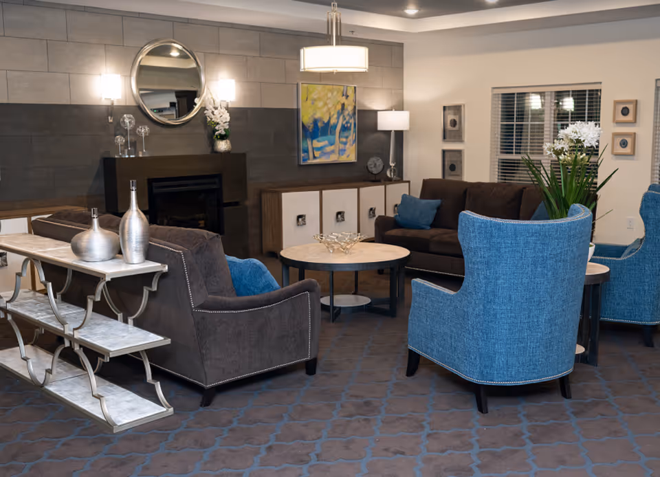 A cozy living room area with brown sofas and blue armchairs arranged around two round coffee tables. The room features a fireplace with a round mirror above it, wall sconces, a floor lamp, and decorative artwork on the walls. There are also decorative vases on a console table behind one of the sofas and a plant near the window with blinds.