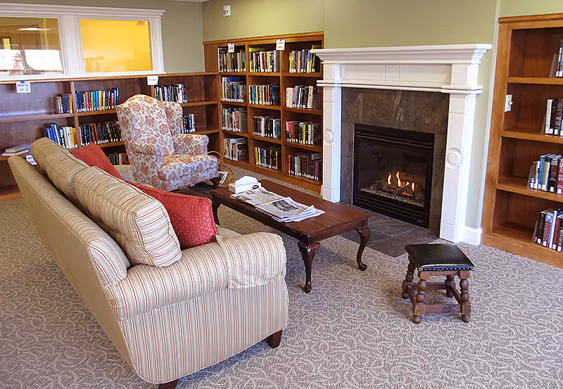 A cozy living room area with a striped sofa, a floral armchair, a wooden coffee table with newspapers on it, a small wooden stool, a lit fireplace with a white mantel, and bookshelves filled with books along the walls.