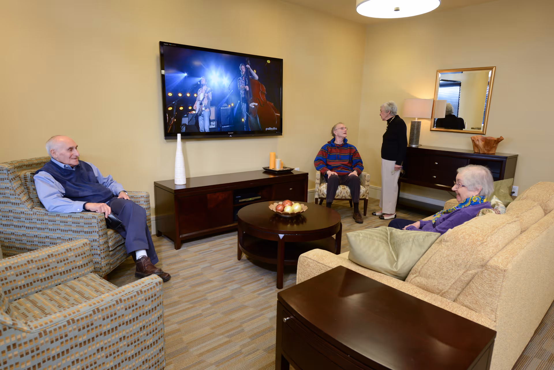 A cozy living room in an independent living facility with four elderly people. Two men are seated in armchairs, one woman is seated on a beige sofa, and another woman is standing and talking to one of the seated men. A flat-screen TV is mounted on the wall showing a musical performance. The room has warm yellow walls, a round wooden coffee table with a bowl of decorative apples, and wooden furniture including a TV stand and a sideboard with a lamp and mirror above it.