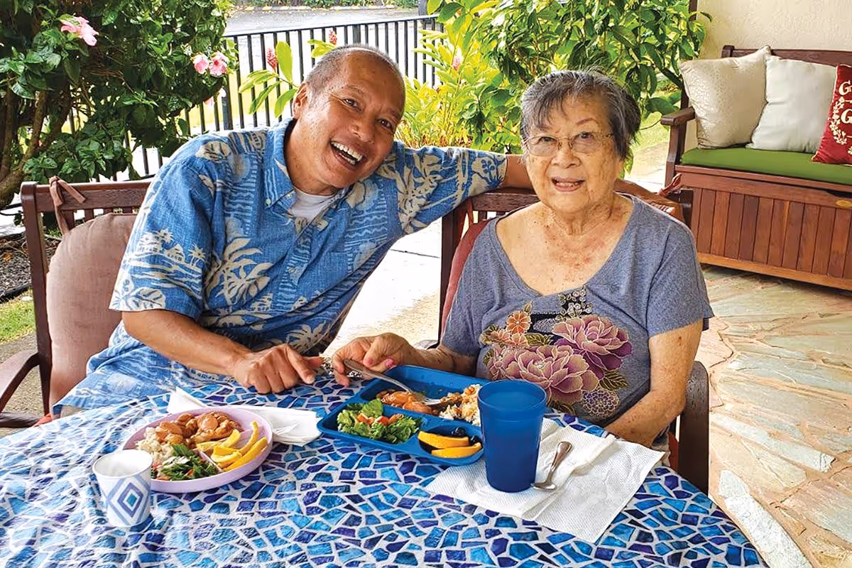 An elderly woman and a middle-aged man sitting at a mosaic-tiled outdoor table enjoying a meal together. The woman is wearing glasses and a floral shirt, while the man is wearing a blue patterned shirt. Behind them is a wooden bench with cushions and green plants.