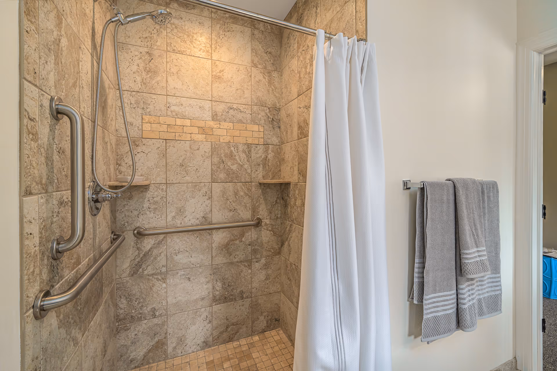 A tiled walk-in shower with beige and brown stone tiles, equipped with a handheld showerhead and multiple stainless steel grab bars. A white shower curtain is partially drawn to the side. On the right wall, there is a towel rack holding three gray towels with white stripes.