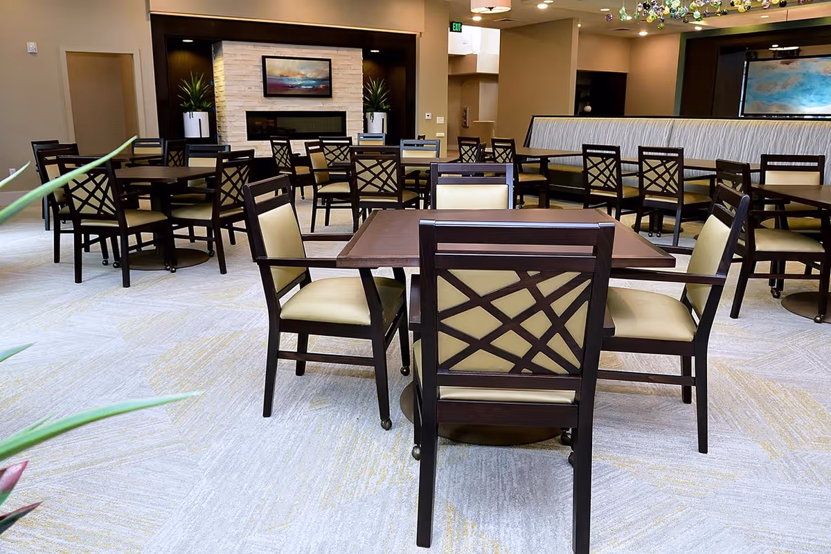 A spacious dining room with multiple dark wood tables and chairs with light beige cushions. The room features a light-colored carpet with subtle patterns, a modern fireplace with a stone surround, two potted plants on either side, and a framed abstract painting above the fireplace. The walls are painted in neutral tones, and there is a long cushioned bench along one wall.